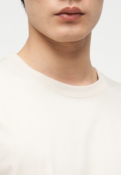 Κοντινό πλάνο στο κάτω μέρος του προσώπου και τον λαιμό ενός ατόμου που φοράει μια απλή λευκή μπλούζα με στρογγυλή λαιμόκοψη, μπροστά σε ουδέτερο φόντο.
