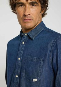 Chemise en denim bleu foncé avec un col boutonné, une poche avant et des boutons clairs. Le tissu a une texture lisse et un design minimaliste.