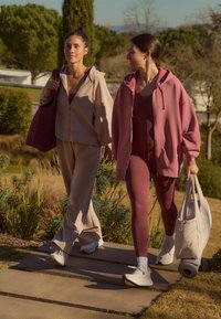 To kvinder går udenfor. Den ene har en lys beige træningsdragt med lynlåshætte på; den anden har en pink oversized hættetrøje og bordeaux leggings.