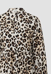 Blouse met luipaardprint en knopen in een lichte stof. Beschikt over een kraag, geplooide taille en zwarte knopen tegen een beige achtergrond.