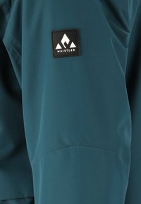 Turquoise waterdichte jas met een gladde textuur, voorzien van een rechthoekige zwarte patch met een wit logo op de linker mouw.