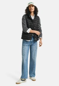 Schwarze Steppweste mit grauen Strickärmeln, kombiniert mit hellblauen weiten Jeans und einem weißen Hemd mit Knöpfen. Beigefarbene Sneakers runden das Outfit ab.