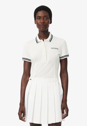 Mujer que lleva una camisa polo blanca de Lacoste con rayas marinas en el cuello y las mangas, combinada con una falda blanca plisada.
