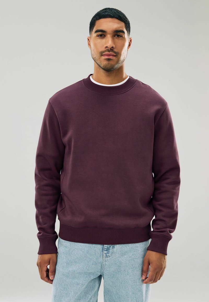 Sudadera burdeos con cuello y puños de canalé. Textura suave; combinada con denim azul claro. Diseño simple y clásico sin patrones.