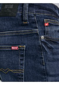 Prim-plan al buzunarului din spate al unei perechi de blugi din denim albastru închis, cu cusături galbene și etichete roșii ale mărcii Big Star.