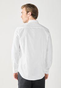 Camicia bianca a maniche lunghe con polsini abbottonati, orlo curvo e tessuto di cotone liscio. Presenta un colletto classico e un design semplice.