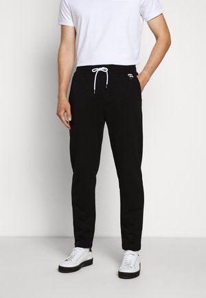 Mand iført sorte snørebukser, hvid T-shirt og hvide sneakers står mod en ensfarvet baggrund.