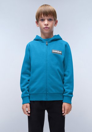 Sudadera azul con cremallera y bolsillo frontal, puños y dobladillo acanalados, con un parche de logo negro en el lado izquierdo del pecho. Pantalones negros usados por debajo.