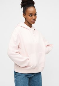 Ανοιχτόροζ φούτερ με κουκούλα κατασκευασμένο από μαλακό υλικό, με μπροστινή τσέπη και κορδόνια. Συνδυάζεται με μπλε τζιν παντελόνι.