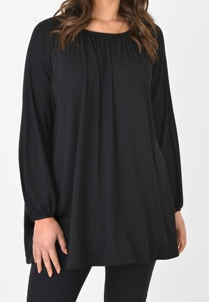 Frau trägt locker sitzende schwarze Tunika mit gerafftem Ausschnitt und schwarze Hose, steht vor einfachem weißen Hintergrund.