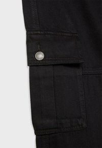 Gros plan sur la manche d'une veste en denim noir avec une lanière boutonnée et des coutures visibles sur le tissu.