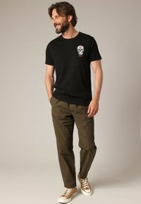 Zwarte t-shirt met korte mouwen met een schedelafbeelding, gecombineerd met olijfgroene broek en tan sneakers. Glad katoen met een ontspannen pasvorm.