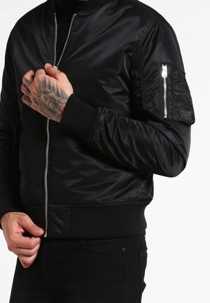 Osoba w czarnej kurtce bombowca z zamkiem błyskawicznym, kieszonką na rękawie na zamek i widocznymi tatuażami na lewej dłoni, poprawiająca kurtkę.