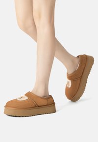 Brown fleece slip-on shoes with a plush accent and a thick tan sole; features a rounded shape and open back for easy wear.