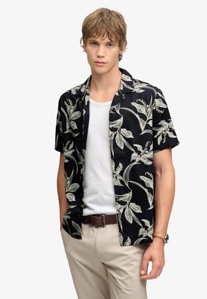 Jeune homme portant une chemise noire à manches courtes avec un imprimé botanique blanc sur un T-shirt blanc, pantalon beige, ceinture marron et montre-bracelet.