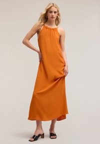 Vestido largo sin mangas de color naranja con escote fruncido, tela suave y corte holgado. Se lleva con unas mule negras de punta abierta, con tacón cuadrado.
