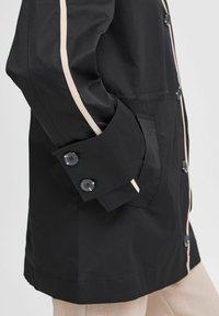 Zwarte waterdichte jas met beige streepdetails, voorzien van een knoopsluiting bij de mouwen, zijzakken en een rechte snit.