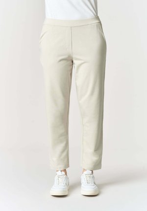Persona con pantaloni beige a gamba stretta e scarpe da ginnastica bianche in piedi contro uno sfondo chiaro e semplice.