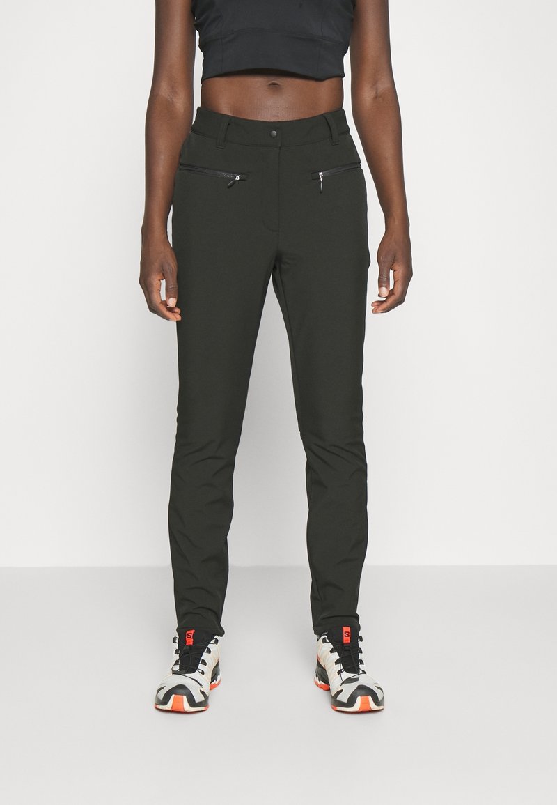 Pantalones ajustados verde oscuro con una textura suave, que presentan bolsillos frontales con cremallera y una cintura clásica, combinados con zapatillas blancas y naranjas.