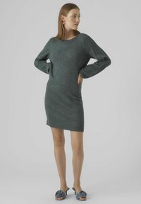 Abito lavorato a maglia verde scuro con scollatura ampia, maniche lunghe e una silhouette leggermente aderente, abbinato a mule grigi con tacco.