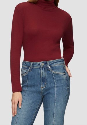 Kobieta ubrana w dopasowany bordowy golf i wysokie niebieskie dżinsy z detalem ozdobnego szwu z przodu, stojąca na jednolitym tle.