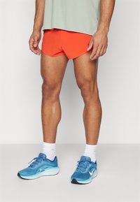 Røde idrettsshorts med en tettsittende design og hvit logo, kombinert med lette blå løpesko og hvite ankelsokker.