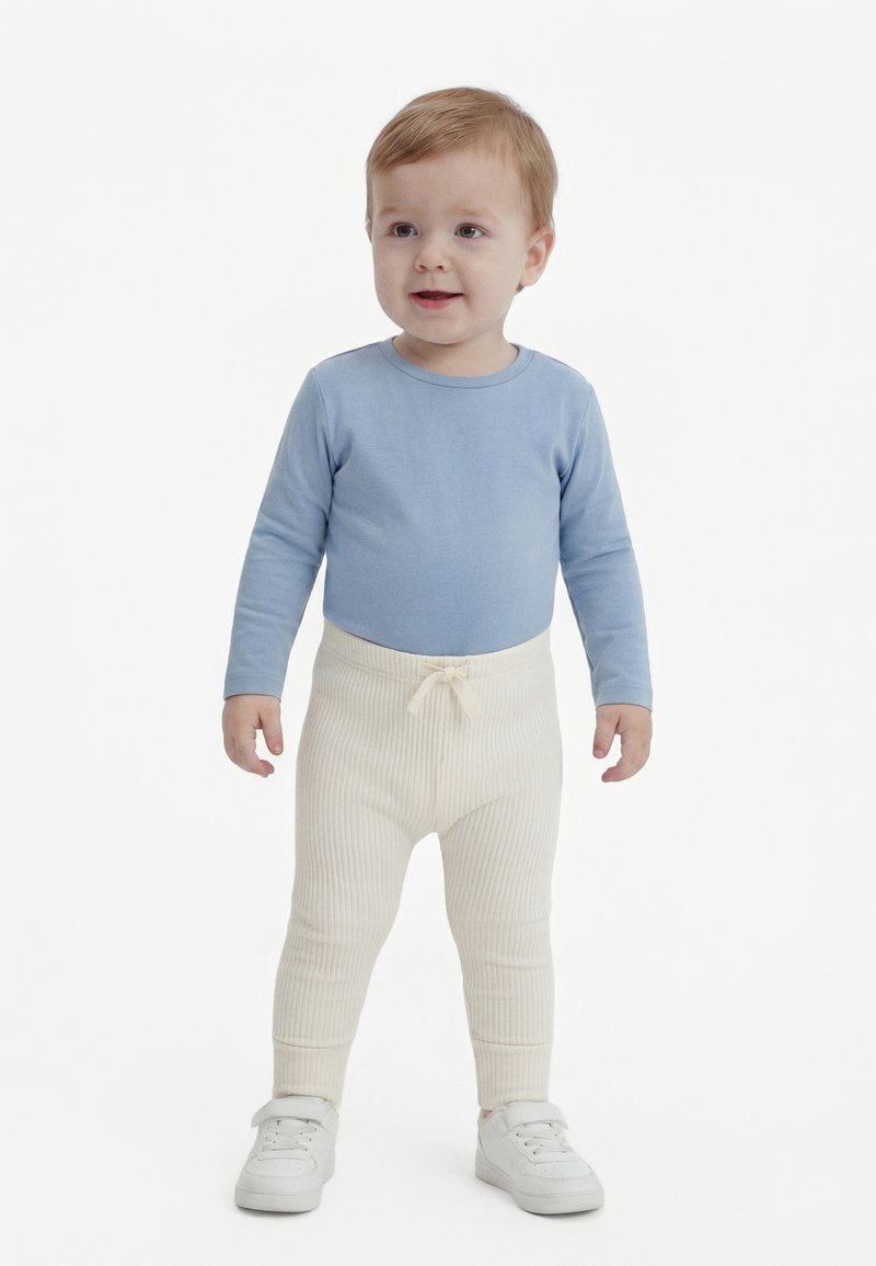 Niño pequeño de pie vestido con una camiseta azul claro de manga larga, pantalones crema acanalados con cordón y zapatillas blancas sobre un fondo blanco.