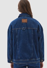 Person med långt brunt hår som bär en mörkblå denimjacka och matchande jeans, vänd bort mot en ljusgrå bakgrund.