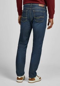 Homme portant un jean bleu foncé et des baskets blanches avec des accents bruns, debout de dos face à la caméra contre un fond uni.