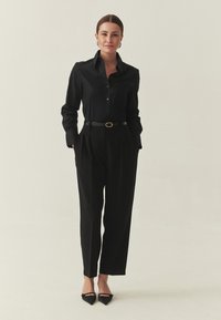 Fekete galléros ing, gombos elülső résszel, magas derekú, pliszírozott fekete nadrággal párosítva. Tartalmaz egy vékony fekete övet és hegyes orrú fekete magas sarkú cipőket.