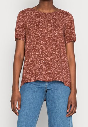 Kobieta ubrana w rdzawy top w kwiaty z krótkim rękawem, w połączeniu z niebieskimi, wysokim stanem dżinsami, stojąca na jednolitym białym tle.