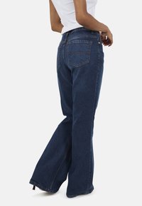 Ciemnoniebieskie dżinsy dzwony z wysokim stanem, z dwoma tylnymi kieszeniami i subtelnymi detalami przeszycia; ścięte na kostce, tworząc szeroką nogawkę.