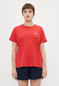 Červené bavlnené tričko s krátkymi rukávmi a okrúhlym výstrihom. Má malú bielu logo FILA na ľavej strane. Skombinované s tmavozelenými šortkami.