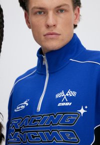 Młody mężczyzna w niebieskiej kurtce wyścigowej na zamek z białym obszyciem i grafiką z szachownicą, stojący przodem z neutralnym wyrazem twarzy.