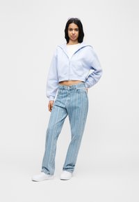 Γυναίκα που φοράει ανοιχτό μωβ κοντό φούτερ, ανοιχτό μπλε ριγέ τζιν, λευκά αθλητικά παπούτσια και γυαλιά ηλίου πάνω στο κεφάλι της, στέκεται μπροστά σε λευκό φόντο.