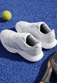 Chaussures de sport blanches sur une surface bleue texturée à côté d'une balle de tennis jaune et d'un manche de raquette de tennis noir.