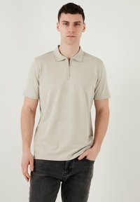 Hellbeige Poloshirt mit kurzem Reißverschlusskragen, aus glattem Stoff gefertigt. Kurze Ärmel, taillierte Silhouette, kombiniert mit dunklen Jeans.