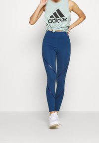 Leggings atletici blu con accenti viola curvi e una fascia alta in vita, abbinati a una canottiera verde chiaro con un logo nero.