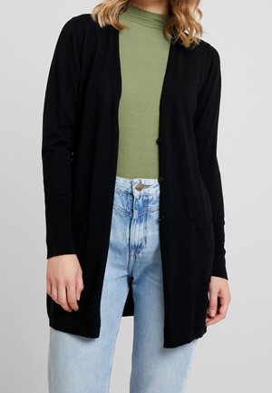 Nainen pukeutuneena mustaan, napitettuun neuletakkiin vihreän resoripaidan ja vaaleansinisten korkeavyötäröisten farkkujen päällä, seisoo yksinkertaisen taustan edessä.