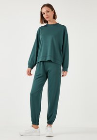 Completo loungewear turchese con felpa a collo rotondo e maniche scese, pantaloni jogger affusolati con polsini elastici, abbinati a sneaker bianche.