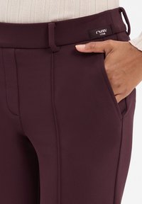 Hand in de zak van donkere paarse op maat gemaakte broek met riemlussen en een klein stoffen etiket nabij de taille, gedragen met een licht geribbelde top.