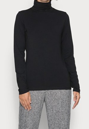 Henkilö pukeutuneena yksiväriseen mustaan pooloneuleeseen ja mustavalkoisiin kuvioituihin housuihin, seisoo kädet rentona sivuilla.