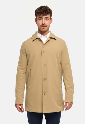 Junger Mann, der einen beigefarbenen, geknöpften Mantel über einem weißen Hemd trägt und vor einem schlichten hellgrauen Hintergrund steht.