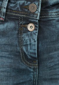 Vaqueros de mezclilla azul oscuro con herrajes de cobre, que presentan botones de latón, costuras en contraste y una sutil textura desgastada cerca de la zona de los bolsillos.