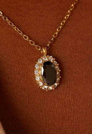 Collier en pendentif en or avec une pierre noire ovale entourée de strass transparents. La chaîne a des maillons allongés, mise en valeur sur un teint de peau marron.