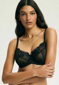 Kobieta z długimi ciemnymi włosami, nosząca czarny koronkowy stanik, spogląda przed siebie, z jedną ręką skrzyżowaną, a drugą opartą na ciele, na jednolitym tle.