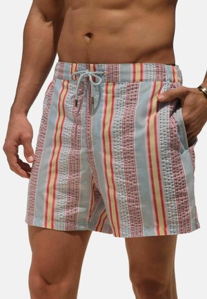 Man wearing pastel-colored, vertically striped swim shorts with drawstring waist and side pockets, hand in pocket, bare torso visible.