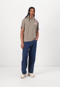 Grå kortermet t-skjorte med logo, kombinert med blå jeans og beige sneakers. Modell som står med hendene i lommene mot en ensfarget bakgrunn.