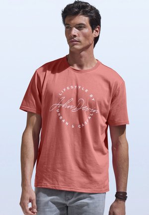 Mann trägt ein korallenfarbenes T-Shirt mit Rundhalsausschnitt und weißem, rundem Schriftzug "Lifestyle by John Devin Urban & Casual" sowie hellgraue Jeans vor einem schlichten Hintergrund.