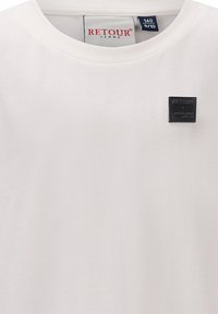 T-shirt bianco in cotone con collo rotondo e un piccolo patch logo quadrato nero che riporta la scritta "RETOUR JEANS". Texture liscia, design semplice.